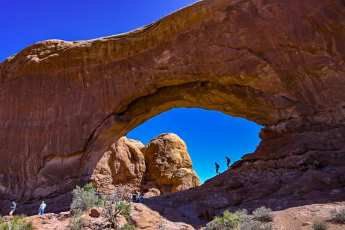 A natural rock arch with people exploring beneath it, surrounded by red rock formations and a clear blue sky.