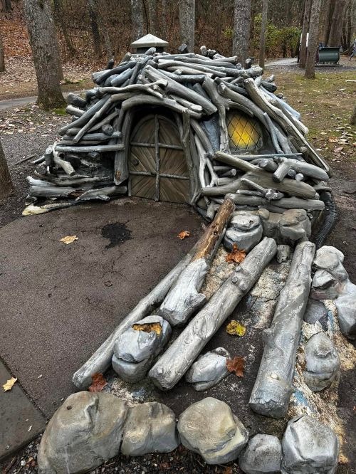 A whimsical playhouse made of logs and stones, resembling a small hut, nestled among trees in a park.