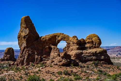 A large rock formation with a natural arch, surrounded by desert landscape and a clear blue sky.