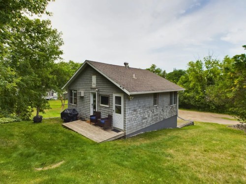 A small gray house with a deck, surrounded by green grass and trees, set against a cloudy sky.