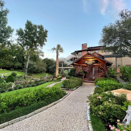 A scenic pathway leads to a wooden entrance of a lodge, surrounded by lush greenery and trees under a clear sky.