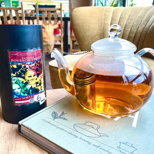 A glass teapot filled with amber tea next to a black tea tin labeled "Vanilla Bourbon Street" on a wooden table.
