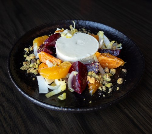 A vibrant dish featuring creamy cheese, colorful citrus, beets, and crushed pistachios on a dark plate.