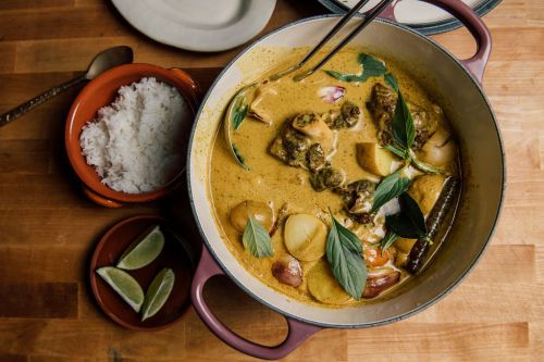 A pot of curry with vegetables and herbs, served with rice and lime wedges on a wooden table.