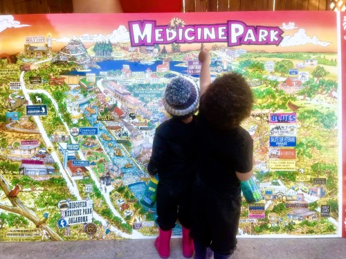 Two children in rain boots point at a colorful map of Medicine Park, surrounded by illustrations of local attractions.