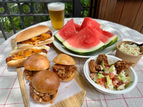 A table with hot dogs, sliders, coleslaw, watermelon slices, and a beer, set outdoors.