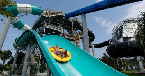 Two people slide down a water slide on a yellow raft, with a large water park structure in the background.