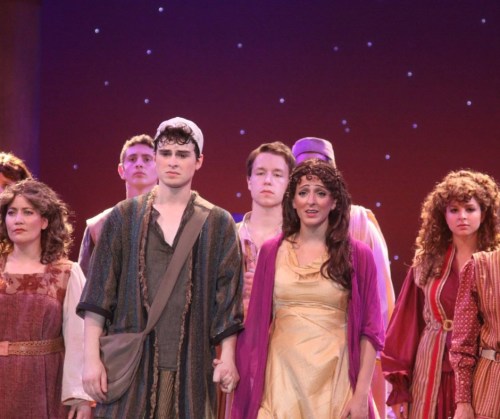 A group of actors in colorful costumes performing on stage, with a focus on two characters in the foreground.