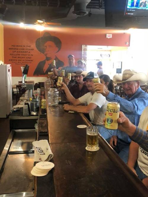 A lively bar scene with patrons raising drinks, a cowboy hat in the foreground, and a mural of a cowboy on the wall.