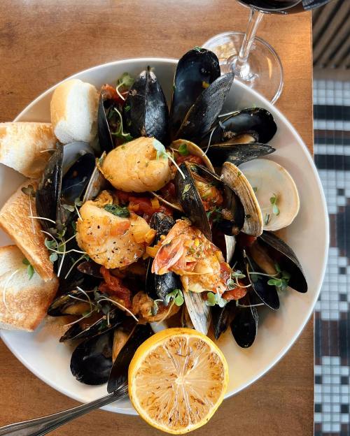 A plate of seafood featuring mussels, clams, shrimp, and lemon, served with toasted bread and a glass of red wine.