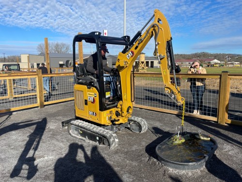 A man operates a yellow CAT excavator, lifting a circular object at a construction site with spectators in the background.