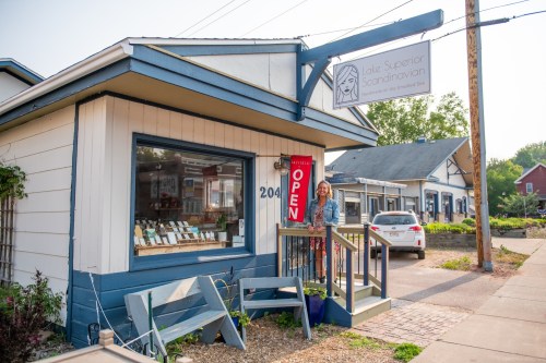 A woman stands outside the Lake Superior Scandinavian shop, which has an "OPEN" sign, on a sunny day.
