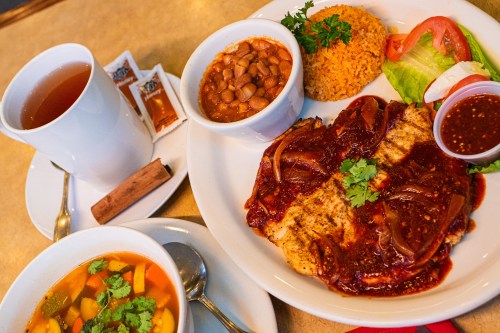 A plate of grilled fish topped with sauce, rice, beans, salad, and a cup of soup, alongside a cup of tea.