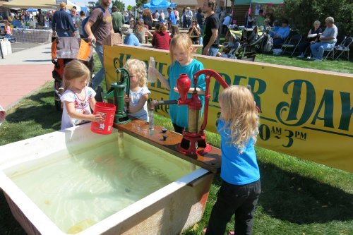 Four children play with water pumps at a community event, filling buckets from a large tub in a sunny outdoor setting.