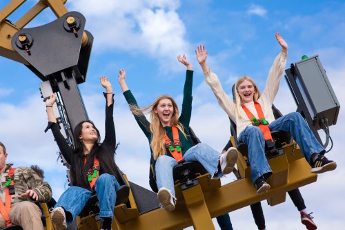 Three young women on a roller coaster, smiling and raising their hands in excitement against a blue sky.