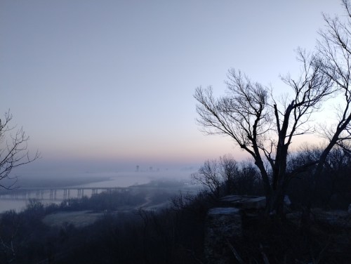 A misty landscape at dawn, featuring bare trees and a river with distant city silhouettes in the background.