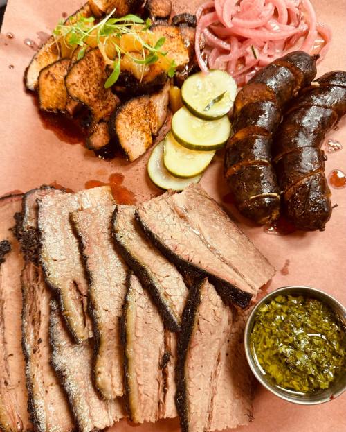 A platter of sliced brisket, smoked sausage, pickles, and pickled onions, garnished with herbs and a green sauce.