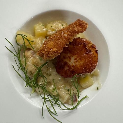 A plate featuring a golden-brown scallop, crispy garnish, and creamy sauce with diced vegetables and herbs.