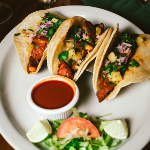 Three tacos filled with meat, pineapple, and herbs, served with a side of salsa and fresh vegetables on a plate.