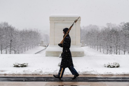 A soldier in winter attire marches with a rifle through falling snow at a memorial site.