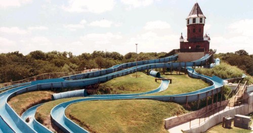A large water slide with multiple twists and turns, leading to a tower structure, surrounded by greenery and blue sky.
