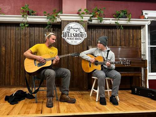 Two musicians play acoustic guitars on stage at Hillsboro Public House, surrounded by wooden decor and plants.
