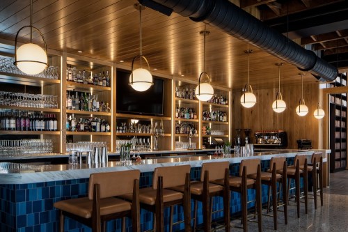 A modern bar with a marble countertop, wooden shelves filled with bottles, and stylish pendant lights overhead.