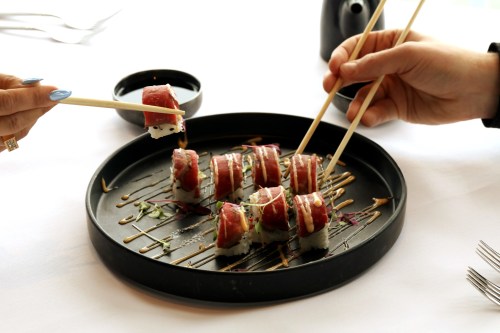 Two hands using chopsticks to pick up sushi rolls on a black plate, with a small bowl in the background.