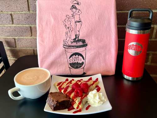 A cozy café scene featuring a latte, dessert crepes with raspberries, a brownie, and branded merchandise on a table.