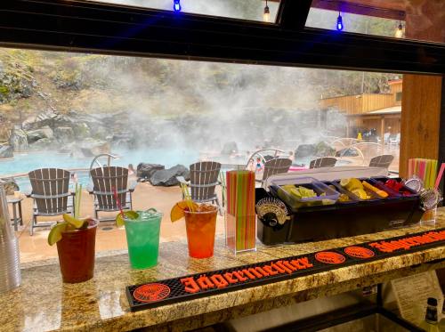 A bar with colorful cocktails in front of a steaming hot spring and lounge chairs in a scenic outdoor setting.