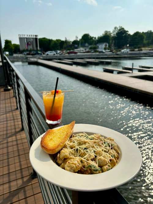 A plate of pasta with garlic bread and a colorful drink, overlooking a marina with boats and a sunny sky.