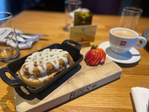 A cinnamon roll drizzled with icing on a wooden board, accompanied by a strawberry and a cup of coffee.