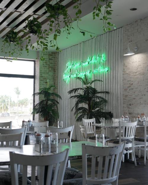 A bright restaurant interior with white tables, green plants, and a neon sign on the wall.
