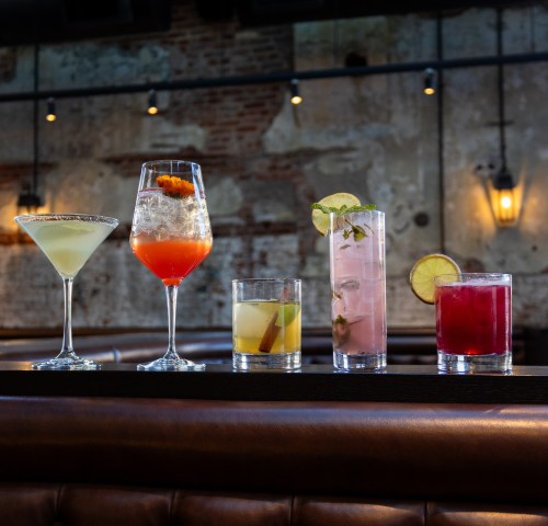 Five colorful cocktails displayed on a bar, featuring various garnishes and glass styles in a stylish setting.