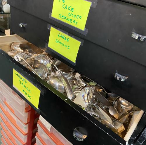 A drawer filled with various silverware, including large spoons and tongs, labeled with bright yellow tags.