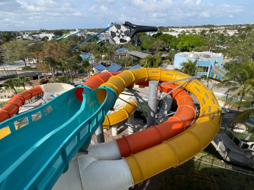 Colorful water slides twist and turn in a sunny amusement park, surrounded by palm trees and vibrant attractions.