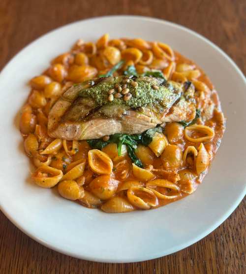 Grilled salmon on a bed of creamy pasta shells with spinach and pine nuts, served on a white plate.