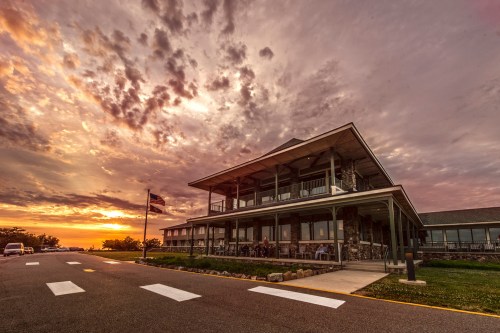 A modern building with a stone facade at sunset, featuring a porch and people sitting outside, under a colorful sky.