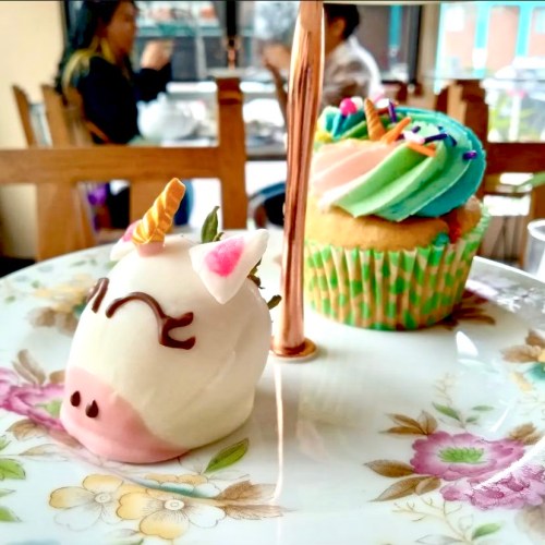 A cute unicorn-shaped dessert and a colorful cupcake on a floral plate, with a cozy café background.