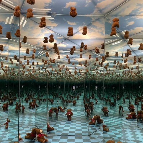 A mirrored room filled with teddy bears, creating a surreal, infinite reflection effect against a blue sky backdrop.