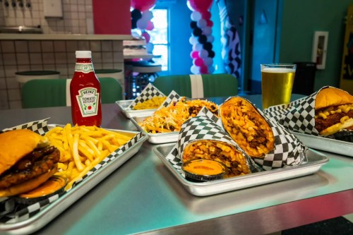 A table with burgers, fries, loaded nachos, and a beer, with ketchup in the background and colorful decor.