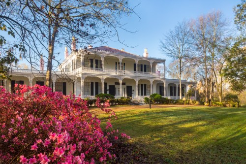 A historic house with a wraparound porch, surrounded by blooming pink flowers and lush green grass under a clear sky.