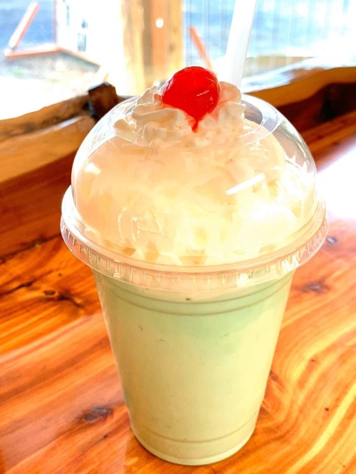 A mint green milkshake topped with whipped cream and a cherry, served in a clear cup on a wooden table.