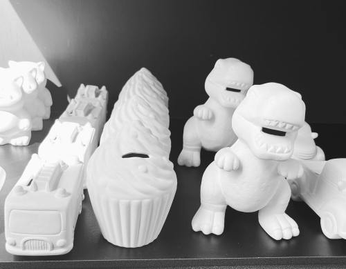 A collection of white ceramic figurines, including dinosaurs, a cupcake, and toy vehicles, displayed on a shelf.