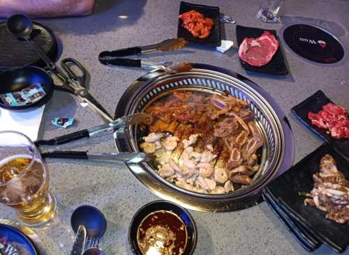 A table set for Korean BBQ with a grill, various meats, side dishes, and a glass of beer.