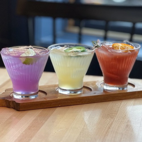 Three colorful cocktails in clear glasses on a wooden tray, featuring purple, yellow, and red drinks with garnishes.
