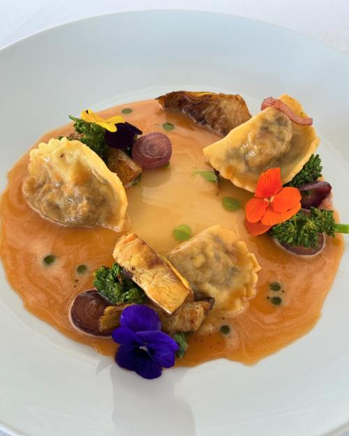 A gourmet dish featuring ravioli in a rich sauce, garnished with edible flowers and vegetables on a white plate.