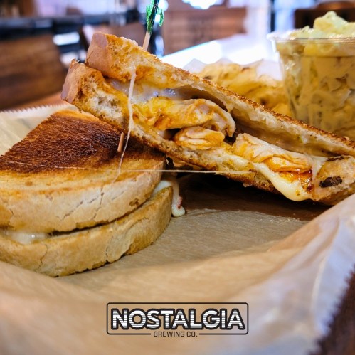 A grilled cheese sandwich with melted cheese and chicken, served with a side of pasta salad. Nostalgia Brewing Co. logo visible.
