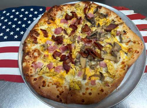 A pizza topped with scrambled eggs, ham, mushrooms, and bacon on a silver tray, set against an American flag background.