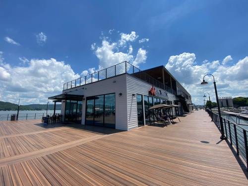A modern waterfront building with large windows, surrounded by a wooden deck and blue sky with fluffy clouds.